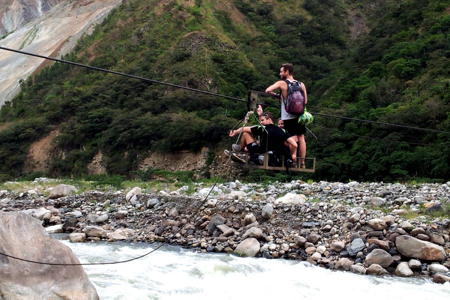Tourists crossing a rushing river inside a rustic steel cable cart called an Oroya in Cusco