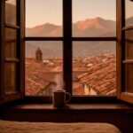 Vista desde la ventana de un alojamiento en Cusco al amanecer, con los tejados de San Blas y una taza de café humeante.