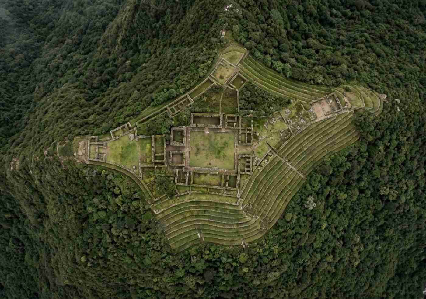 Vista aérea de dron del sitio arqueológico de Choquequirao, mostrando la plaza principal y las terrazas circundantes ubicadas en una silla de montaña rodeada de densa vegetación de bosque nuboso.