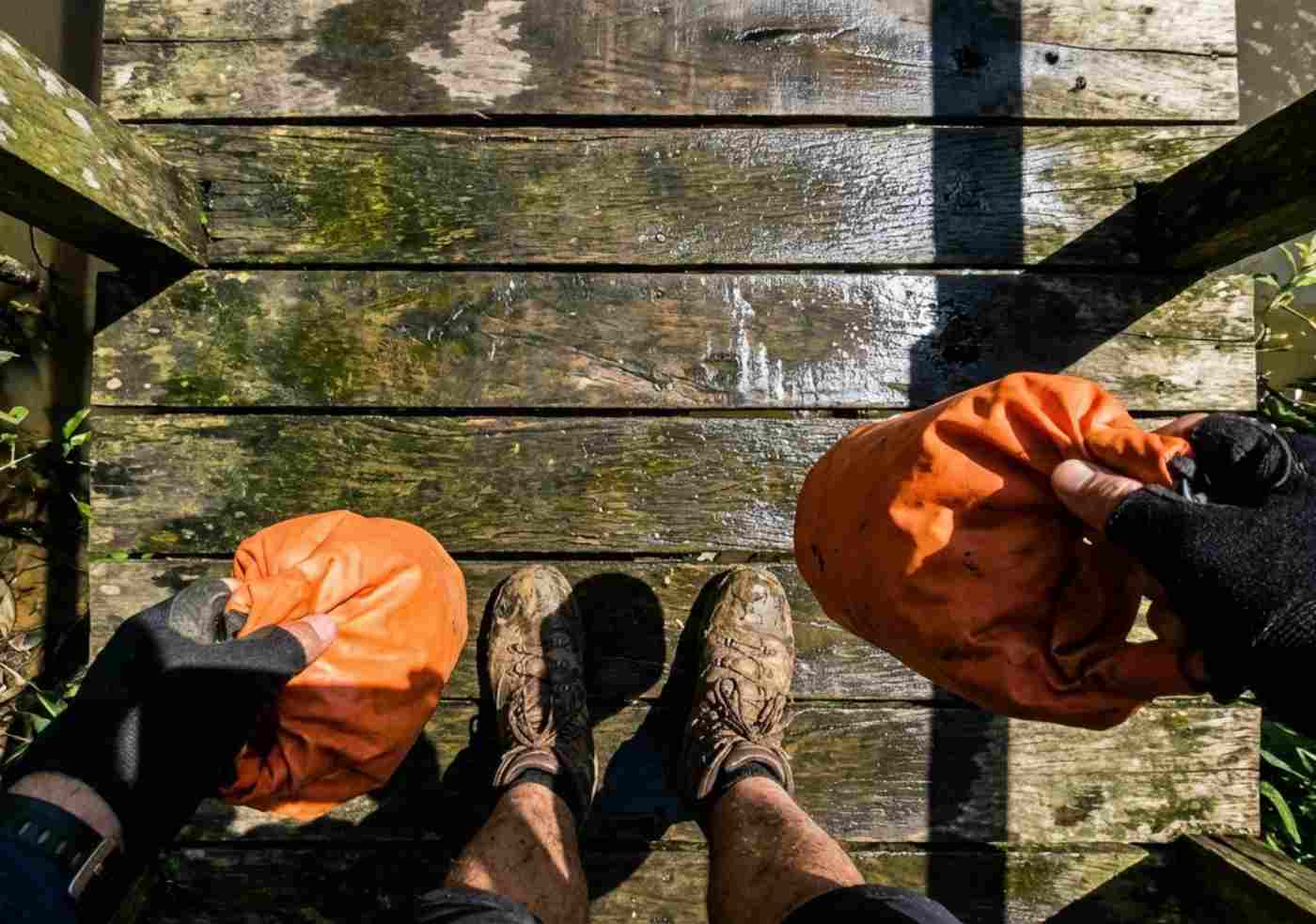 Perspectiva en primera persona (POV) de unas botas de trekking con barro sobre un muelle de madera rústico en Iquitos, sosteniendo una mochila estanca.