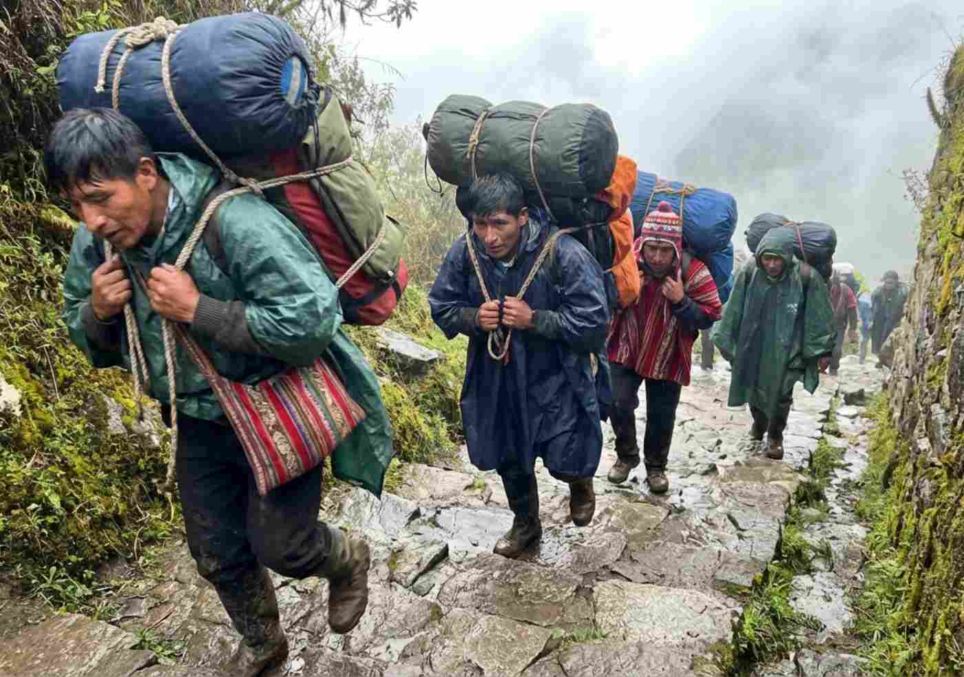 Un grupo de porteadores quechuas cargando grandes bultos y equipo de campamento subiendo por un sendero empinado y nebuloso del Camino Inca.
