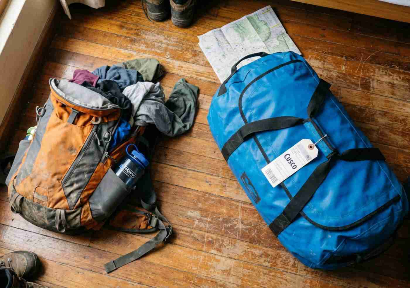 Foto casual de una mochila pequeña abierta y un bolso grande impermeable sobre un piso de madera rústico en un hostal de Cusco, mostrando el proceso de empaque.