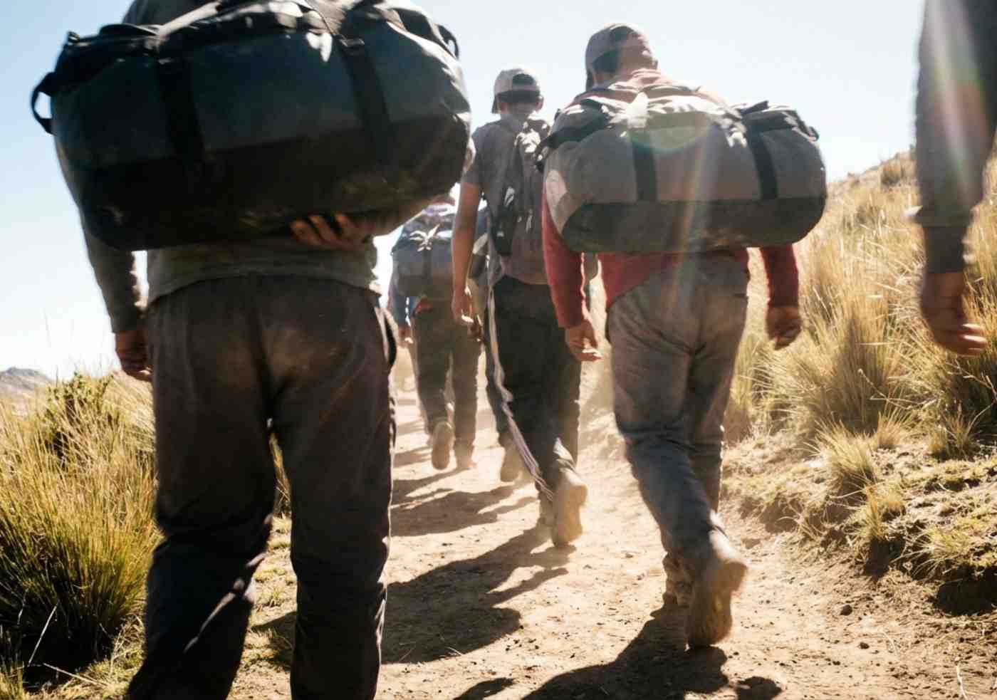 Fila de porteadores quechuas con uniformes y duffels grandes caminando por un sendero de tierra estrecho, vista desde atrás.