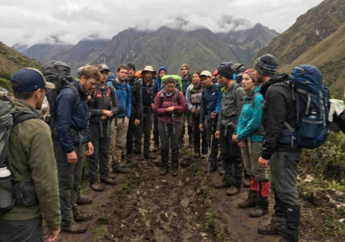 Fotografía de estilo documental tomada con celular que muestra a un grupo numeroso de unos 15 senderistas con mochilas grandes caminando en fila por un sendero estrecho y embarrado en los Andes peruanos. El clima está nublado y se ve cansancio en sus posturas. Refleja la experiencia de un tour grupal económico.