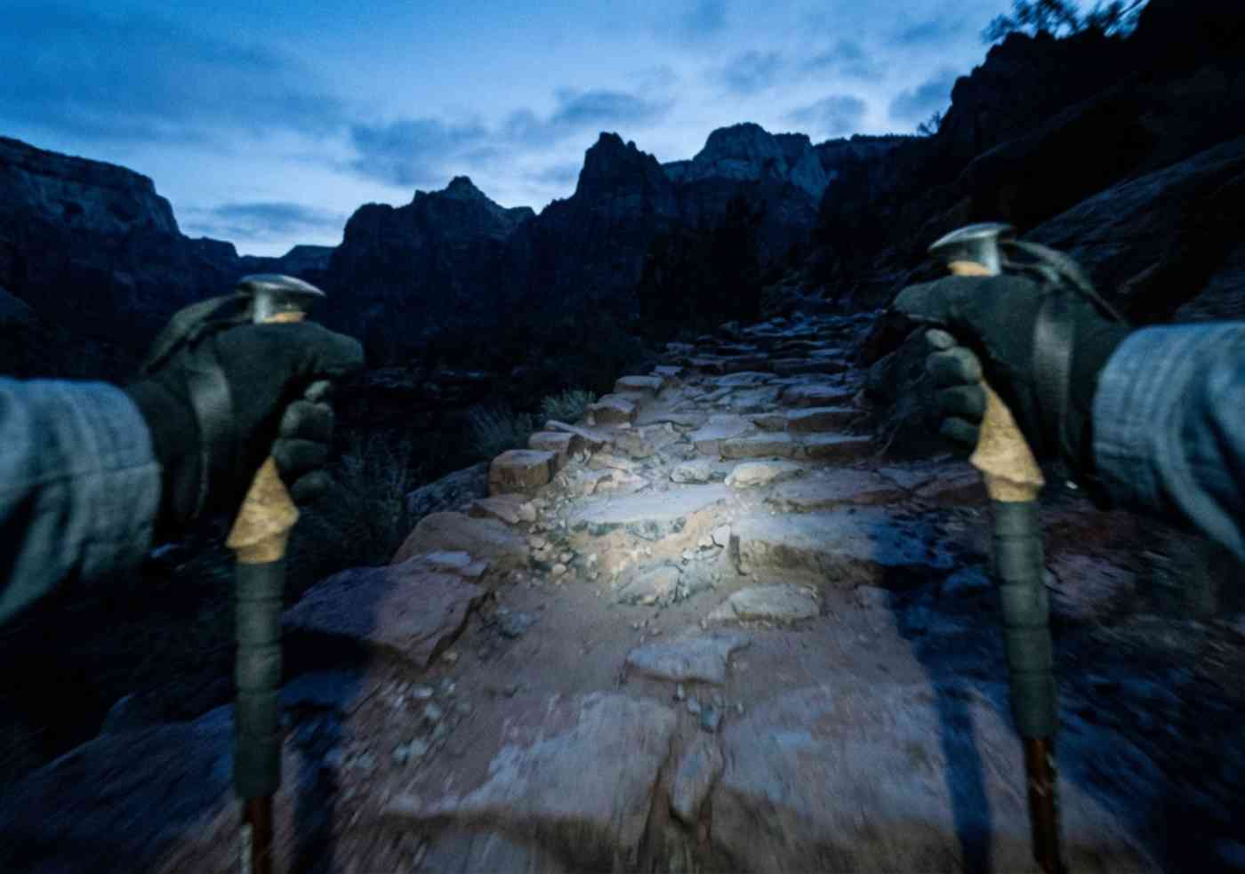 Perspectiva en primera persona (POV) de un senderista usando bastones en un camino de piedra iluminado por una lámpara frontal al amanecer.