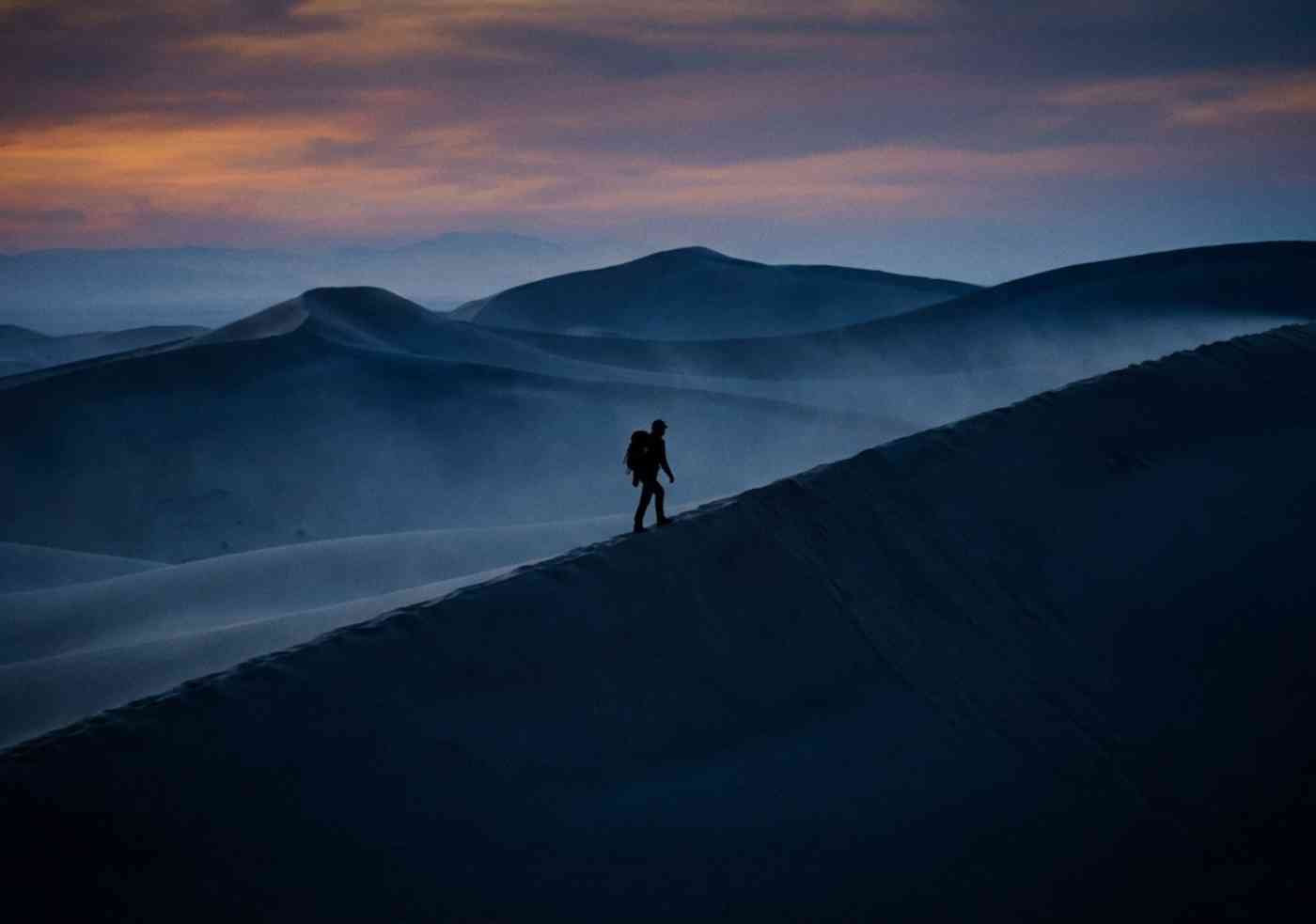 Silueta de un viajero caminando por la cresta de una duna gigante bajo un cielo anaranjado de atardecer en Huacachina.