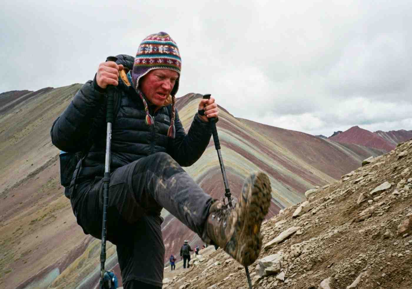 Foto de estilo snapshot de un excursionista con bastones subiendo el sendero empinado hacia la cumbre de la Montaña Arcoíris Vinicunca, mostrando el esfuerzo y el paisaje montañoso.
