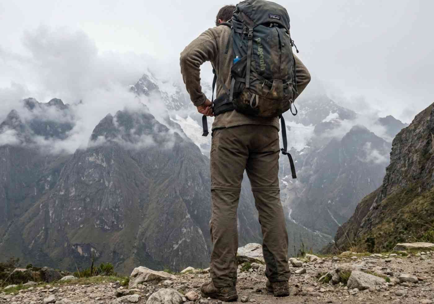 Foto estilo móvil de una persona de espaldas ajustando una mochila de trekking grande y cargada, parada en un sendero de tierra con montañas al fondo.