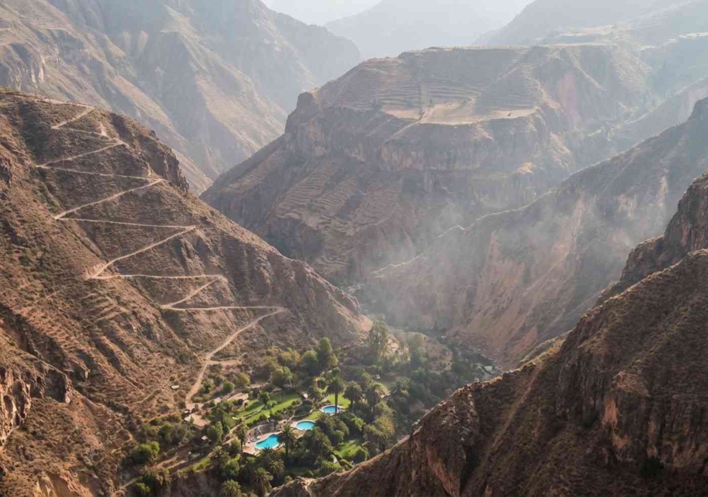 Vista aérea del Oasis de Sangalle con piscinas azules y palmeras en el fondo del árido Cañón del Colca, mostrando el sendero en zigzag que sube por la pared de la montaña.