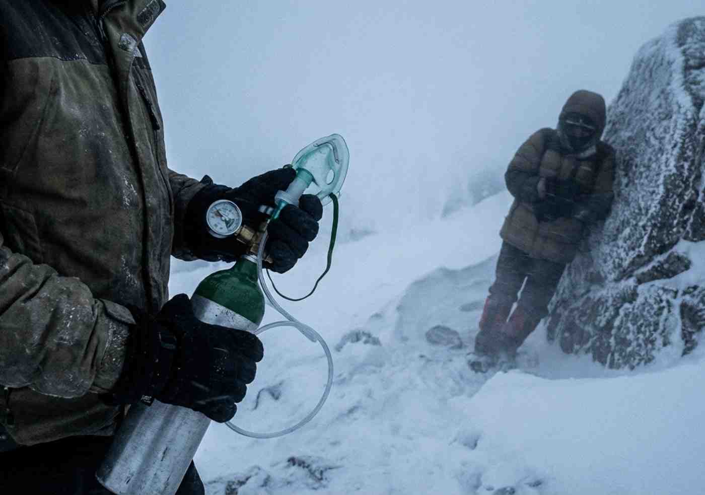 Un guía de montaña revisa un balón de oxígeno medicinal portátil en un paso nevado y con niebla, con un senderista descansando al fondo.