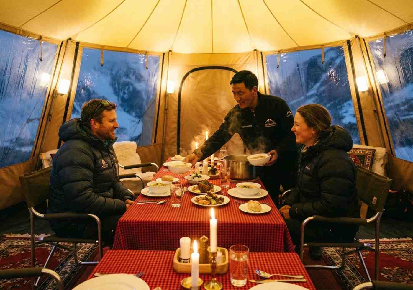 Interior de una carpa comedor bien iluminada y cálida a gran altitud, con un cocinero sirviendo comida caliente a dos senderistas sentados en una mesa.