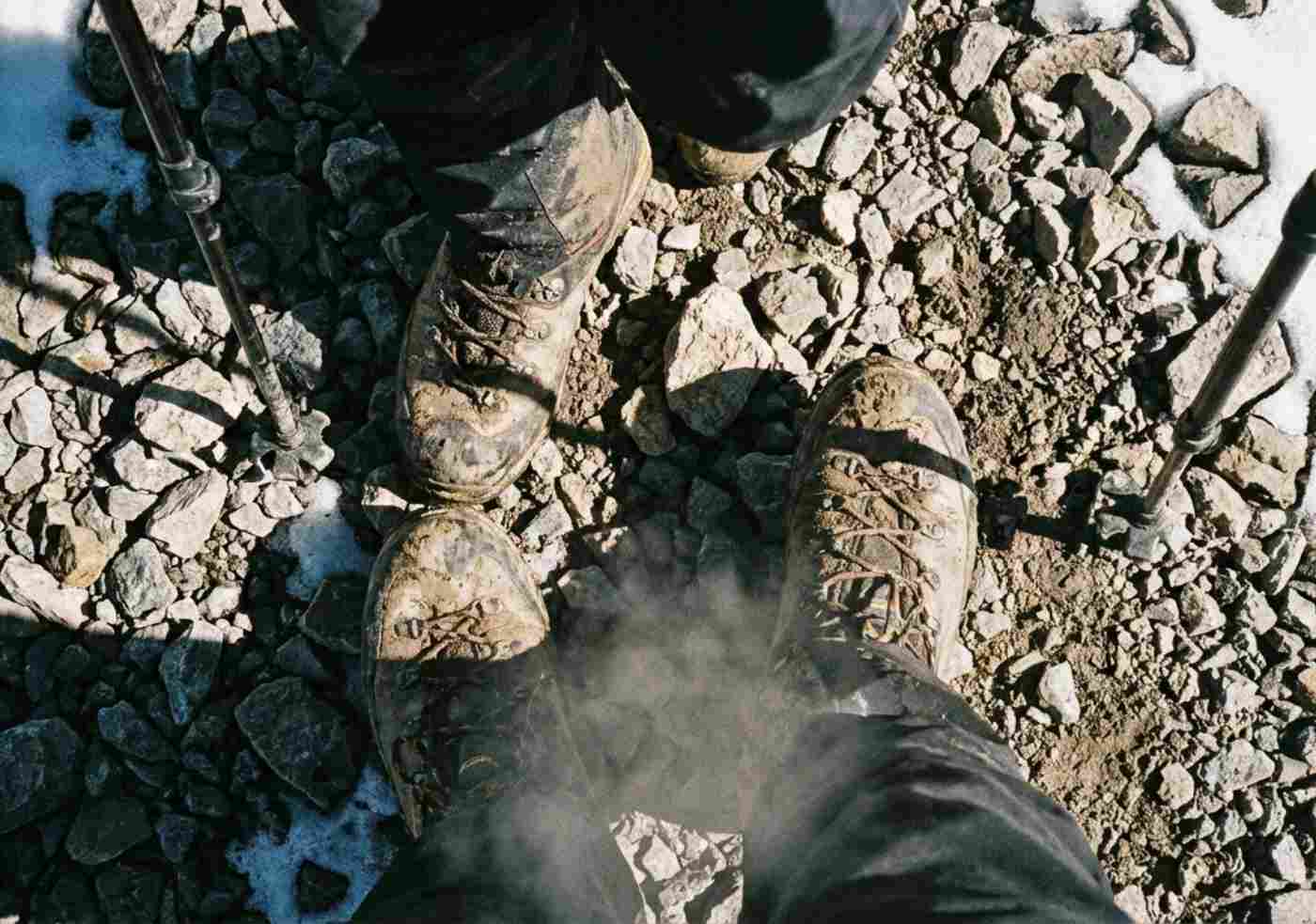 Vista en primera persona mirando hacia abajo a unas botas de trekking cubiertas de barro y polvo pisando rocas sueltas y afiladas en un sendero de alta montaña empinado.